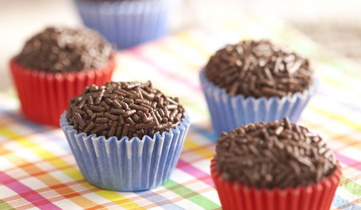 Fotografia em tons de azul e vermelho de uma grade com guardanapo quadriculado branco, verde, rosa, amarelo e azul com cinco brigadeiros e forminhas vermelhas e outras azuis. Ao fundo dois copos, um laranja e um azul. Fotografia em tons de azul e vermelho de uma grade com guardanapo quadriculado branco, verde, rosa, amarelo e azul com cinco brigadeiros e forminhas vermelhas e outras azuis. Ao fundo dois copos, um laranja e um azul.