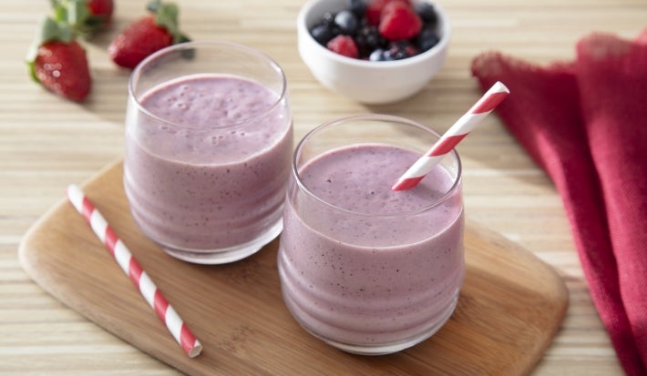 Fotografia de dois copos de vidro pequenos com o smoothie, em um deles têm um canudo vermelho e branco, o outro canudo está ao lado dos copos sobre uma mini tábua de madeira decorativa. A tábua está sobre uma mesa de madeira clara.