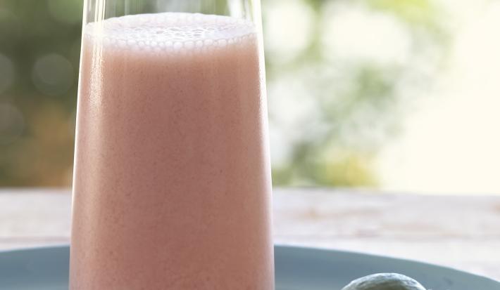 Fotografia de uma vitamina de goiaba com leite e neston com uma fatia fina de uma rodela de fruta na borda do copo de vidro. A bebida está apoiada em um prato azul, sobre uma mesa de madeira.