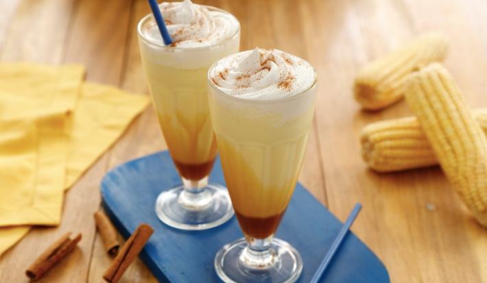 Fotografia em tons de amarelo, em uma bancada de madeira de cor marrom. Ao centro, uma tábua azul contendo 2 copos de milkshake. Ao lado, algumas canelas em pau espalhadas e ao fundo, um pano amarelo e algumas espigas de milho espalhadas.