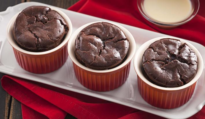 Fotografia em tons de vermelho em uma bancada de madeira escura, uma toalha vermelha, um recipiente branco retangular com três ramequins vermelhos com os suflês de chocolates dentro deles.