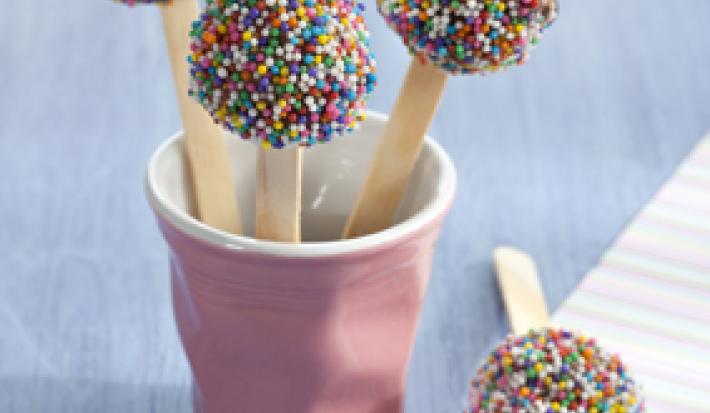 Fotografia em tons de lilás e azul, com copo lilás contendo três pirulitos de brigadeiro com confeitos coloridos, com mais um pirulito ao lado sobre guardanapo colorido, tudo sobre bancada em tons de azul.