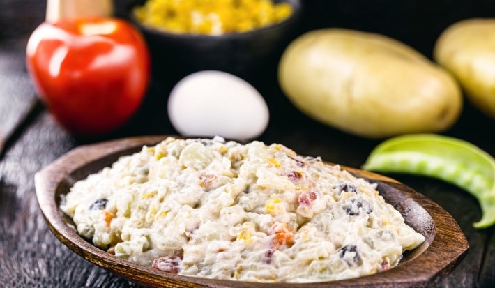 Fotografia de uma salada mista em uma tigela de madeira, com milho, cenoura, uva-passa e um toque de azeitonas, apresentando uma textura cremosa. Ao fundo, um tomate e uma cebola sugerem ingredientes adicionais.