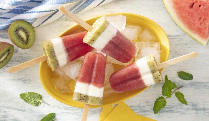 Foto da receita de Picolé de Melancia. Observa-se um prato amarelo fundo com 4 picolés dentro com algumas pedras de gelo. O picolé tem 3 fases: a de baixo, verde, a do meio, uma faixa branca, e a de cima e maior, vermelha, assim como a melancia.
