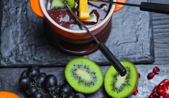 Fotografia de um Fondue de chocolate meio amargo dentro de um recipiente de vidro na cor laranja, com frutas sobre o creme. Ao lado, algumas frutas sobre uma mesa preta, como uvas roxas e um kiwi cortado na metade.