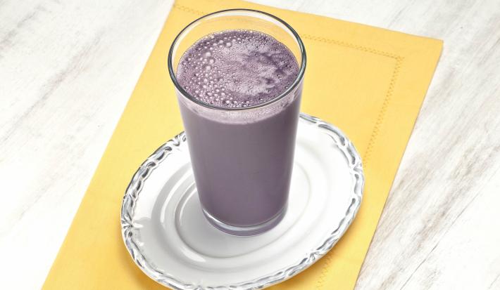 Fotografia em tons de roxo e amarelo em uma bancada de madeira clara, um pano amarelo, um prato branco redondo com um copo de vidro alto e dentro dele a bebida de uva com abacaxi.