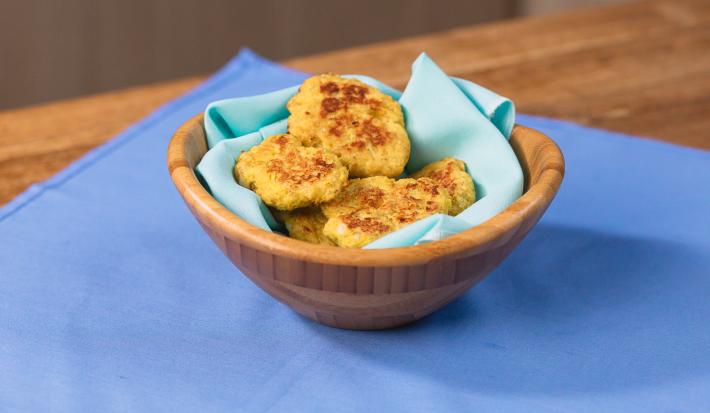 Fotografia em tons claros de uma mesa de madeira com um pano azul. Sobre o pano tem um recipiente de madeira fundo com um paninho azul claro com alguns nuggets saudáveis.