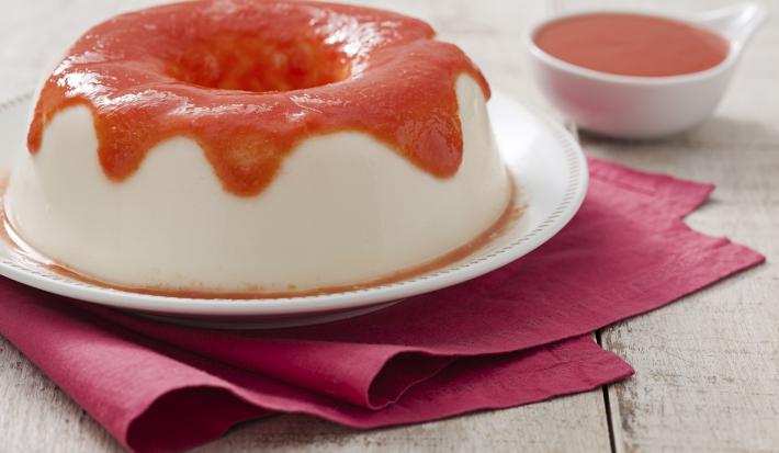 Fotografia em tons de branco, vermelho e cinza, com prato branco ao centro contendo flan coberto com calda vermelha sobre guardanapo bordeaux, ao lado potinho com calda vermelha, tudo sobre bancada na cor cinza.