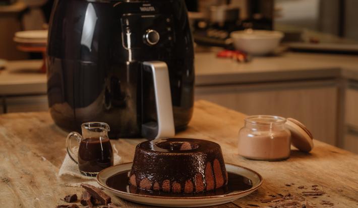 Imagem de um bolo de chocolate com calda de chocolate em um prato. Ao fundo, uma air fryer e ao lado uma jarra pequena com calda.