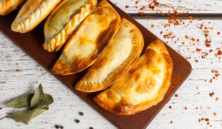 Fotografia vista de cima de cinco pasteis assados na air fryer sobre uma tábua retangular de madeira pequena, a qual está sobre uma mesa de madeira branca.