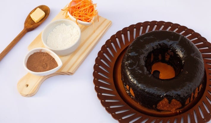 Um bolo de cenoura inteiro, com cobertura de chocolate brilhante cobrindo toda a superfície. Ao lado, há uma tábua com ingredientes usados na receita: manteiga, farinha, cacau em pó e cenoura ralada.