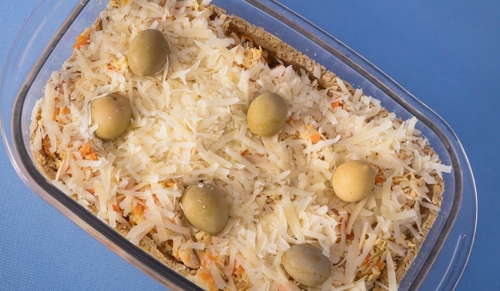 Fotografia vista de cima de um recipiente refratário retangular, de vidro, com uma receita de torta com massa de grão de bico e ascenda, finalizada com queijo parmesão e azeitonas