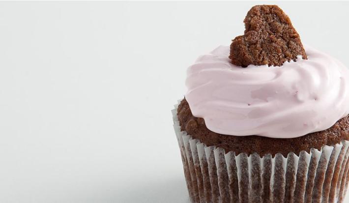 Fotografia em tons de branco, marrom e rosa vista de frente. Contém um bolinho de chocolate com creme de cereja e para decorar um mini bolinho em formato de coração por cima.
