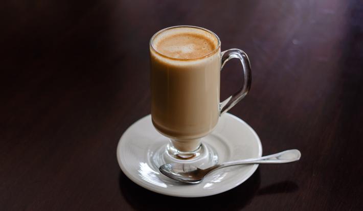 fotografia em tons de preto, branco e marrom de uma bancada preta. Contém um pratinho redondo branco com uma xicara transparente com chocolate cremoso.