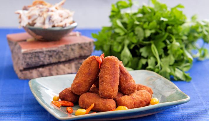 Foto da receita de croquete de carne, servidos em um prato acinzentado, sobre uma bancada azul com um maço de salsinha ao fundo