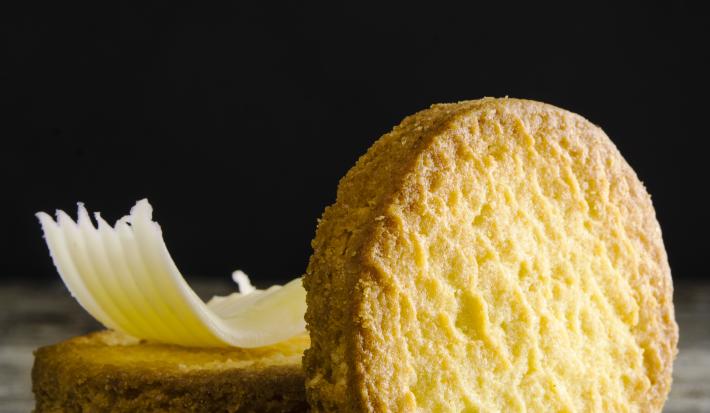 Fotografia de um biscoito em pé apoiado em outros dois biscoitos que estão atrás, e em cima desses que estão empilhados, tem uma lasca pequena de queijo. Estão sobre uma mesa de madeira em tons de cinza.