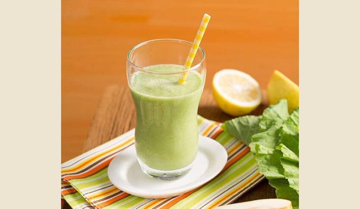 Fotografia em tons de verde e laranja em uma mesa de madeira com um pano listrado colorido e um copo de vidro com o suco verde de pera, couve e gengibre em cima dele. Ao lado, uma laranja cortada ao meio e folhas de couve.