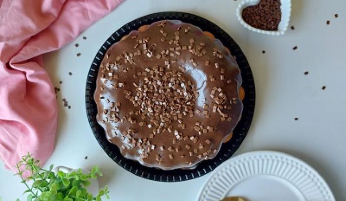 Foto em tons de marrom da receita do melhor bolo de cenoura servido sobre uma base preta com bastante cobertura de brigadeiro por cima com um paninho cor de rosa ao lado