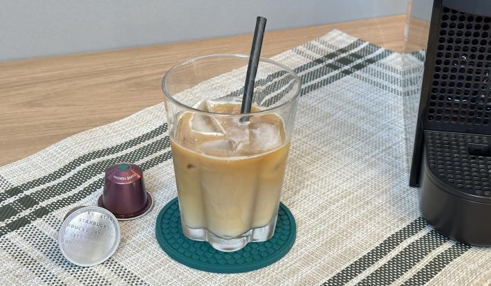 Fotografia em tons de verde em uma bancada de madeira, um copo com o café e cápsulas de starbuclks ao lado.