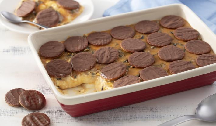 Fotografia em tons de azul em uma bancada de madeira clara, um pano azul, um recipiente retangular vermelho com o pavê de doce de leite, ameixas e biscoito calipso dentro dele. Ao fundo, um prato branco com um pedaço do pavê.