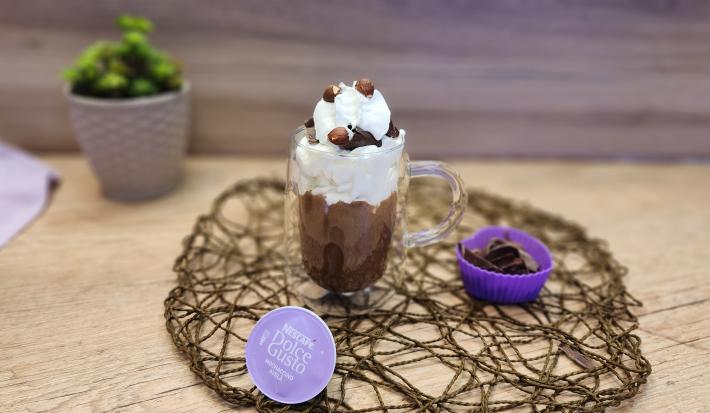 Fotografia em tons de lilás em uma bancada de madeira, com uma xícara alta de vidro e o milkshake de avelã com chocolate dentro dela. Em destaque, a cápsula de Mochaccino Avelã de Dolce Gusto e ao fundo um paninho lilás e um vasinho lilás com planta.