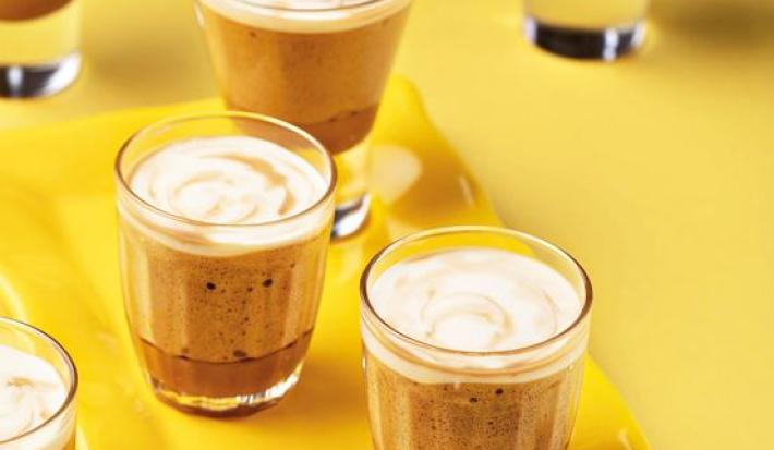 Fotografia em tons de amarelo em uma bancada de madeira amarela, um recipiente amarelo retangular e três copinhos de vidro com a mousse de café e caramelo dentro deles.