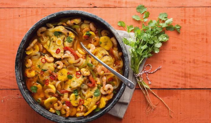 Fotografia em tons de vermelho em uma bancada de madeira vermelha com um recipiente redondo e fundo de pedra com a moqueca de peixe e camarão dentro dela.