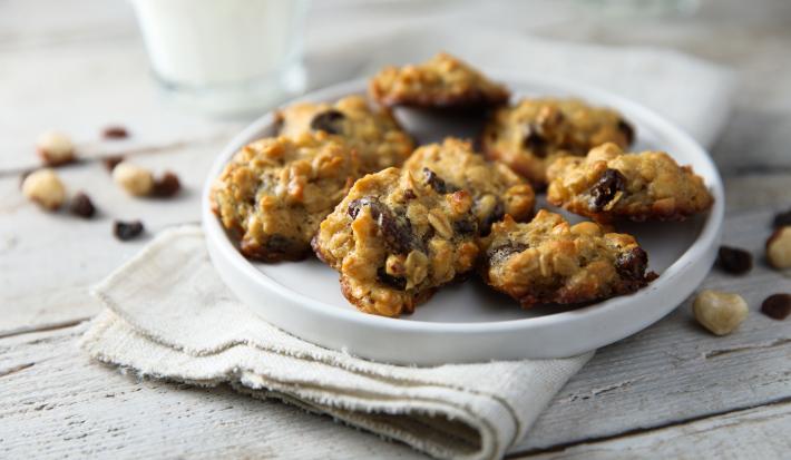 Fotografia em tons de branco, no centro, um prato branco com seis cookies com passas e grãos. Embaixo dos cookies, um pano de prato branco que está sobre uma mesa de madeira na cor branca.
