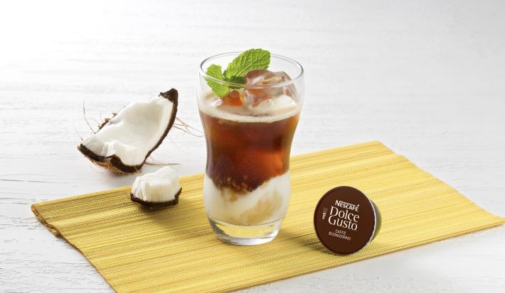 fotografia em tons de cinza, bege e marrom tirada de frente. Contém um tecido marrom e por cima um copo transparente com a bebida, um cubo de gelo e uma folha de hortelã dentro. Ao lado esquerdo uma capsula de Dolce Gusto e ao lado direito fatias de coco