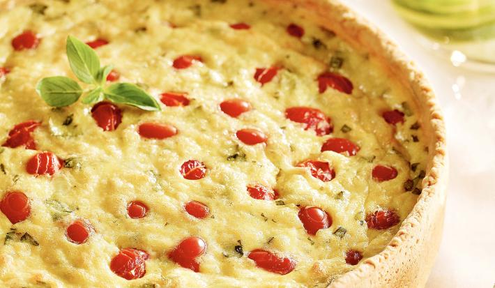 Fotografia em tons de amarelo e vermelho em uma bancada de madeira com uma toalha bege. Ao centro, o quiche de tomates com manjericão.