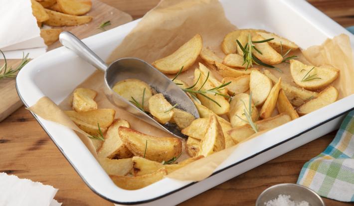 Fotografia em tons de amarelo e verde em uma bancada de madeira clara, um recipiente retangular com batatas feitas no forno e ramos de alecrim em cima dela. Ao lado, um pano xadrez em amarelo e verde.