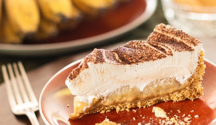 Em cima de uma mesa, fatia de banoffe com massa crocante, doce de leite e chantilly, decorada com canela em pó.