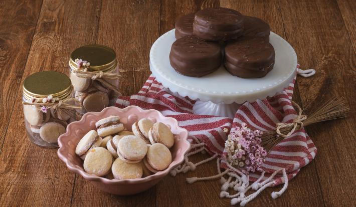 Foto da receita de alfajor com doce de leite, em diversos formatos redondos de tamanhos menor e maior, servidos sobre uma bancada de madeira decora com um tecido vermelho e branco listrado