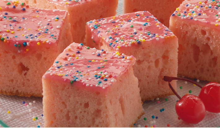 Vários pedaços de bolo rosa cortados em quadrados com confeitos coloridos por cima e duas cerejas ao lado.