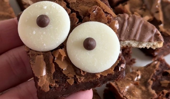 Fotografia de uma mão masculina segurando o brownie de morcego. Ao fundo tem mais pedaços do brownie sobre um prato raso branco.