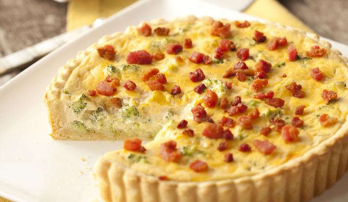 Fotografia em tons de marrom, branco e amarelo de uma bancada de madeira com um paninho amarelo, sobre ele um prato quadrado com um quiche e uma espátula. Ao fundo um prato quadrado branco com uma fatia de quiche.