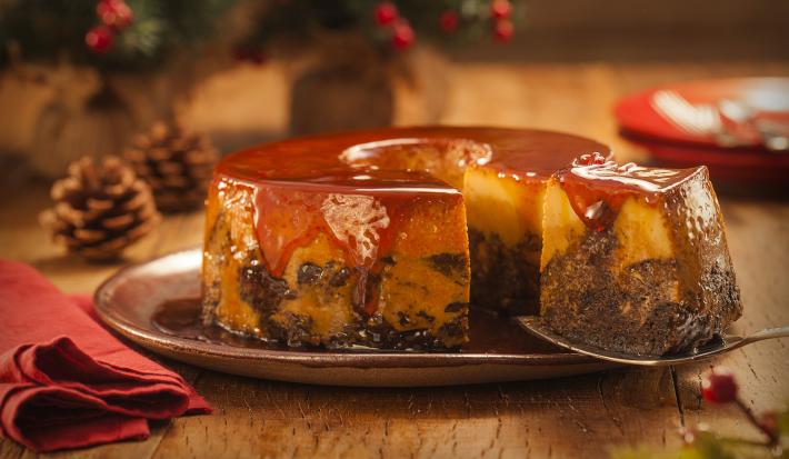 Foto aproximada da receita de Pudim de Panettone com uma fatia sendo retirada. Ele está sobre um prato marrom, numa bancada de madeira decorada com itens de natal