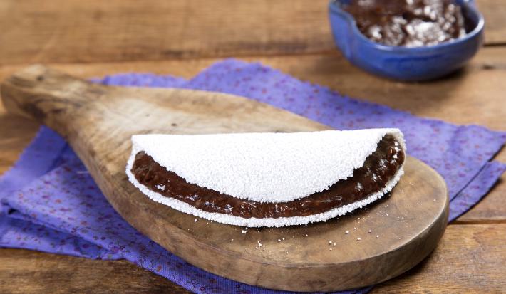Fotografia em tons de marrom e roxo de uma bancada de madeira com um paninho roxo, sobre ele um tábua de madeira com uma tapioca. Ao fundo um recipiente roxo com creme de dark chocolate.