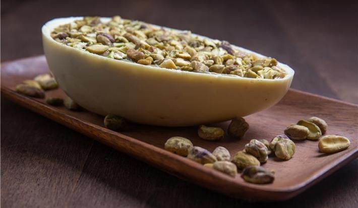 Fotografia de uma metade de ovo de páscoa sobre um apoio retangular de madeira, com alguns pistaches espalhados ao lado. O ovo de páscoa tem a casca de chocoalte branco, o recheio de brigadeiro de pistache e pistaches por cima.
