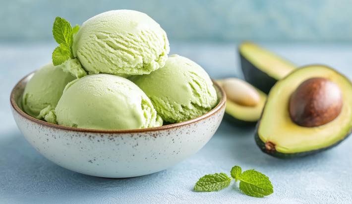 Imagem de um bowl claro com quatro bolas de sorvete de abacate decorado com uma folha de hortelã. Ao lado, abacates cortados ao meio.