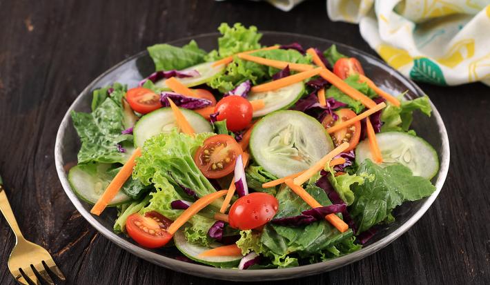 Fotografia de uma salada de folhas, pepino, cenoura ralada, tomates cereja e repolho roxo. Ao lado, um garfo na cor dourado, e ao fundo, uma tolha de mesa dobrada na cor branca, com desenhos em amarelo e verde, sobre uma mesa de madeira na cor preta.