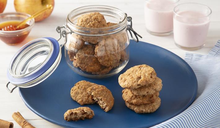 Foto da receita de biscoitos de maçã com iogurte nesfit servida em diversas porções, algumas dentro de um pote de vidro hermético e outras sobre um prato azul escuro com dois copos de iogurte nesfit ao fundo além de um vidro de mel