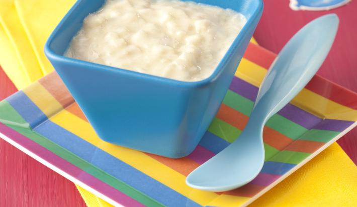 Fotografia em tons de rosa e amarelo em uma mesa de madeira rosa com um paninho amarelo, um prato listrado colorido com um potinho azul em cima dele com o creminho de frutas dentro. Ao lado, uma colher infantil azul.