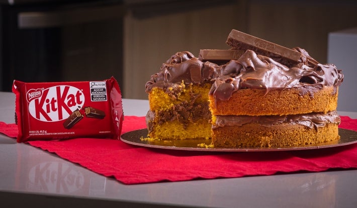Fotografia em tons escuros de um bolo de cenoura com ganache de kitkat no recheio e na cobertura. As laterais do bolo estão sem a ganache, o bolo está apoiado em um pano vermelho sobre uma mesa cinza. Ao lado tem uma embalagem de kitkat.