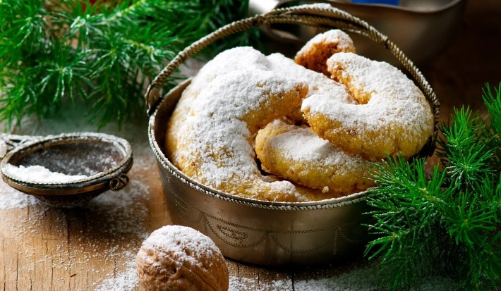 Em cima de uma mesa decorada com enfeites de natal, recipiente com biscoitos em forma de meia lua, polvilhados com açúcar de confeiteiro.