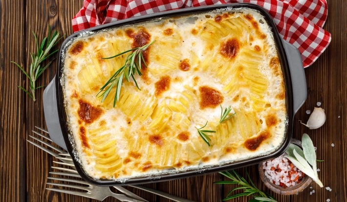 Em cima de uma mesa, refratário com batatas gratinadas com molho branco, decorado com alecrim.