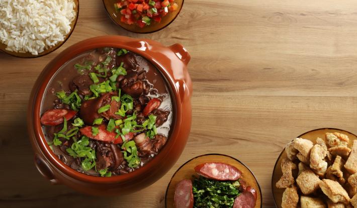 Fotografia vista de cima de uma feijoada completa, com cebolinha por cima, em um recipiente grande clássico de vidro marrom. Ao redor tem acompanhamentos de arroz, torresmo, vingrete e couve, todos em um recipiente de vidro, sobre uma mesa de madeira