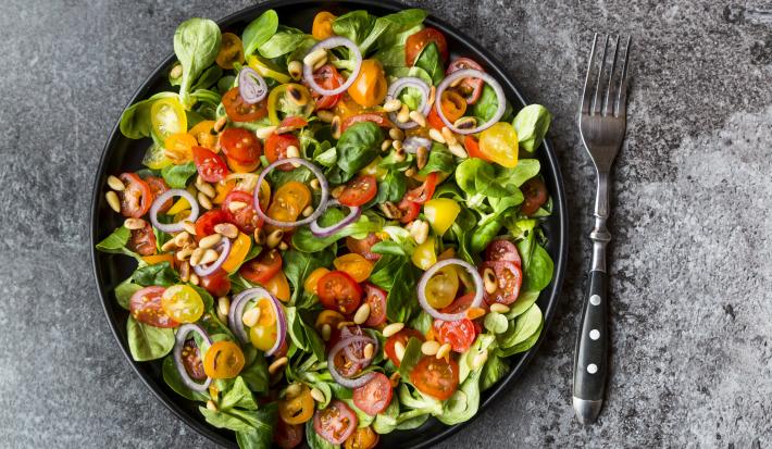 Fotografia vista de cima de uma salada de milho e pequi com folhas, tomates cereja, cebola roxa e grãos em um prato raso preto. Ao lado da salada, um garfo com cabo de cor preta, sobre uma mesa cinza.