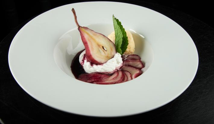 Fotografia de uma sobremesa gelada de pera cozida no vinho com um pouco de creme de leite por cima, juntamente de uma bola de sorvete de creme e uma folha de hortelã. A sobremesa está dentro de um prato branco com o meio fundo.