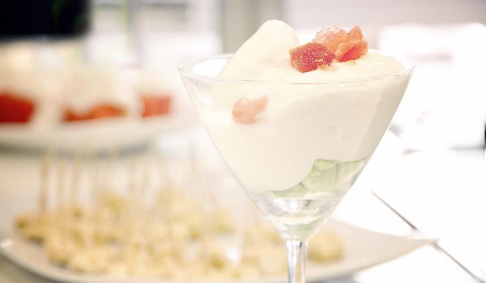 Fotografia de um sorvete de creme com champanhe e raspas de gelo em uma taça. Ao fundo, aperitivos em um recipiente de vidro na cor branco. A granita está sobre uma mesa de madeira na cor branca.
